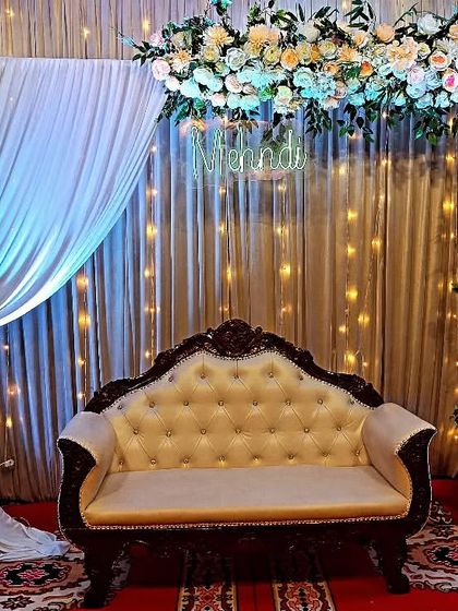 An elegant Mehndi stage with a floral arch, a "Mehndi" neon sign, and shimmering curtains with fairy lights. The traditional sofa and side vases add a touch of class to the setup.