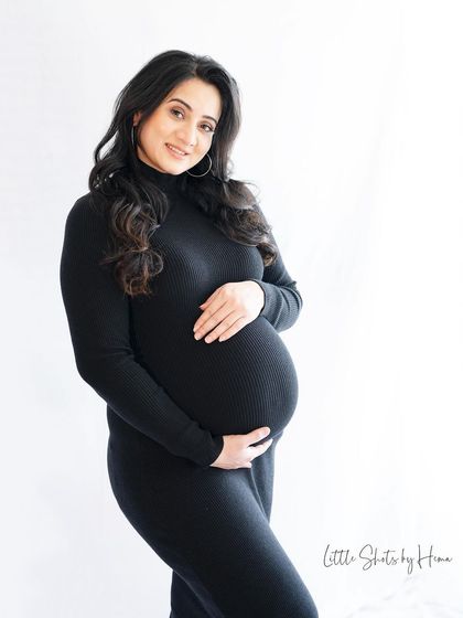 A classic and simple portrait of a mother-to-be in a form-fitting black turtleneck dress. She stands against a clean white background, her hands gently holding her bump.