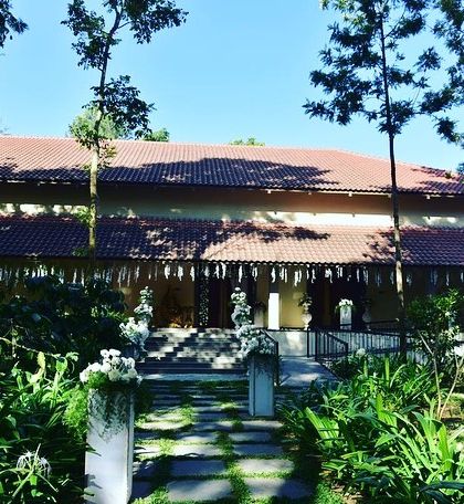 The exterior of a beautiful home decorated for an engagement ceremony. The traditional roofline is accented with simple, elegant hangings, and the pathway is lined with floral arrangements.