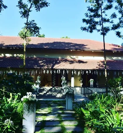 The exterior of a beautiful home decorated for an engagement ceremony. The traditional roofline is accented with simple, elegant hangings, and the pathway is lined with floral arrangements.