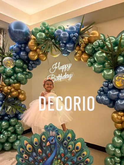 The birthday girl dressed in white, posing in front of her stunning peacock-themed decoration. The rich colors of the blue, green, and gold balloon arch create a beautiful contrast and a perfect photo memory.