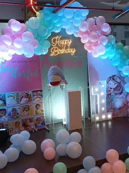 This little mermaid theme for a first birthday uses a blue and pink balloon arch to create an under-the-sea feel. The setup also includes a photo display board and a shark plushie as a fun prop.