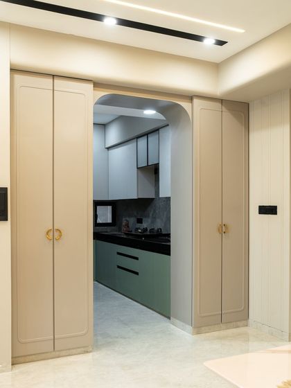 This elegant kitchen entryway blends functionality with soft luxury. The muted taupe cabinetry with rounded edge detailing subtly frames the arched opening, leading into a kitchen that balances bold sage green with calm, light grey textures.