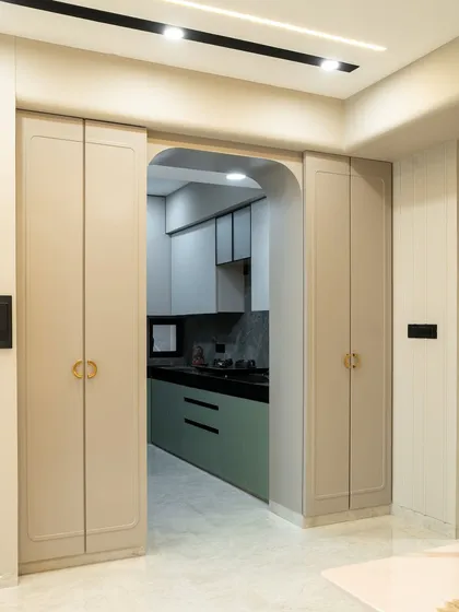 This elegant kitchen entryway blends functionality with soft luxury. The muted taupe cabinetry with rounded edge detailing subtly frames the arched opening, leading into a kitchen that balances bold sage green with calm, light grey textures.