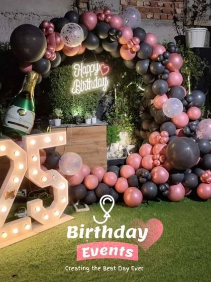 A stylish outdoor setup for a 25th birthday on a lawn. The decor features a pink and black balloon arch, a neon sign, and large marquee numbers.