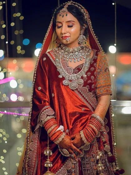 A full-body portrait of a bride in a velvet red lehenga, with beautiful background bokeh from the evening lights.