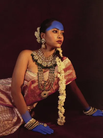 A graceful, reclining pose that showcases the full bridal look, from the layered necklaces to the flowers in her hair. The lighting is soft and painterly.