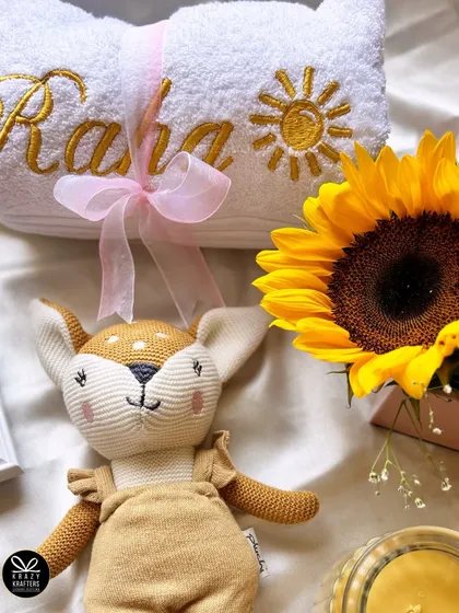 A close-up of the personalized 'Raha' towel with a sun embroidered on it, next to the adorable deer plushie and a bright sunflower.