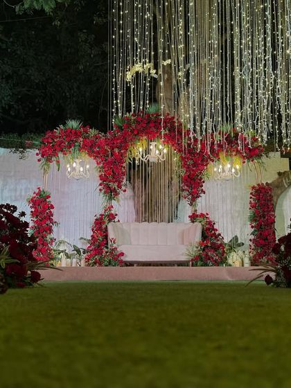 A bold and romantic stage setup for a reception or sangeet. Rich red flowers, elegant chandeliers, and a curtain of lights create a dramatic and glamorous atmosphere.