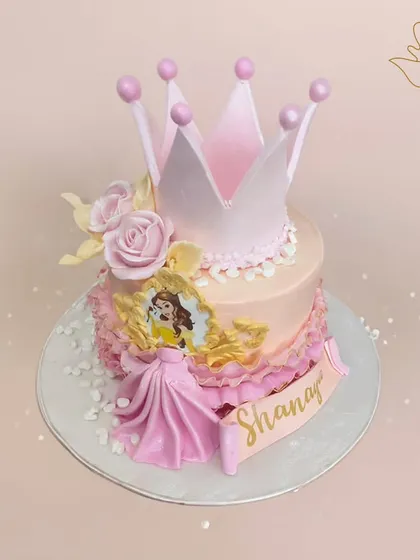 A beautiful Belle-themed princess cake, featuring a golden frame with her portrait, a soft pink crown, and delicate ruffles. It's a tale as old as time, baked into a sweet reality.