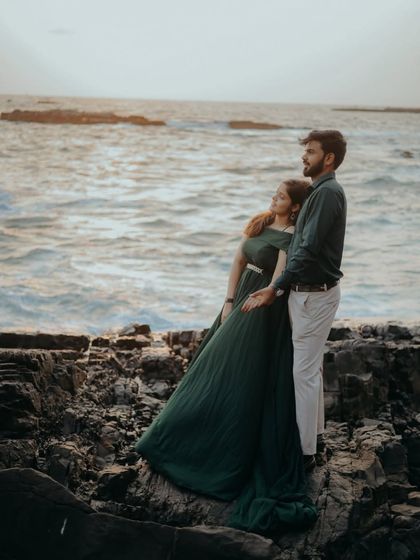 A moody and romantic pre-wedding photo on the rocky shore. The deep green gown flows beautifully, mirroring the movement of the ocean waves.