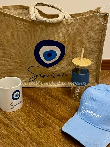 All things evil eye. This coordinated set includes a personalized jute tote bag, a coffee mug, a glass tumbler, and a baseball cap, all featuring the protective symbol.