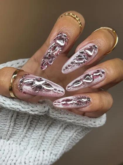 For the ultimate romantic maximalist. These long stiletto nails feature a sheer glitter base adorned with sculpted pink chrome hearts and textures, creating a stunning, high-impact look.