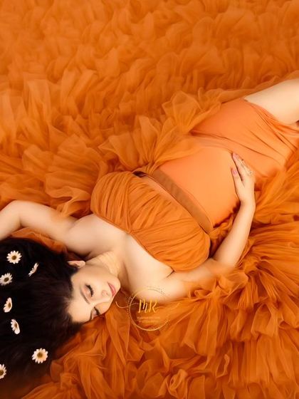 A creative flat-lay style portrait. The mother-to-be lies on a bed of orange ruffled fabric, her hair adorned with small white flowers for an artistic touch.