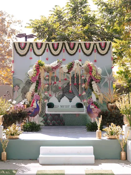 A beautiful Haldi seating arrangement with a custom peacock-themed backdrop. The traditional swing is adorned with lush floral arrangements, blending classic motifs with a fresh, modern aesthetic.