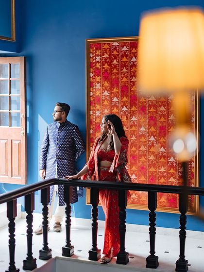 An editorial-style portrait of a couple inside a palace, with a vibrant blue wall and traditional Indian textile art.
