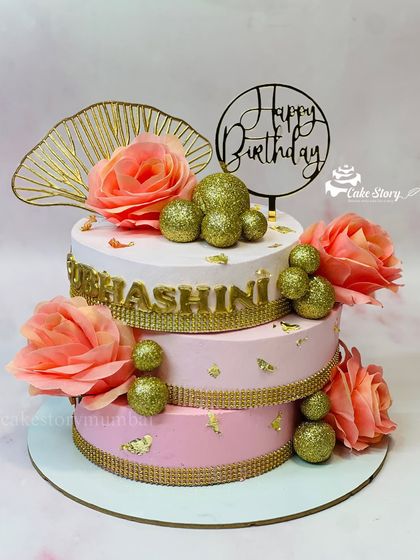 An elegant three-tier cake in shades of pink and gold. This crisscross design is adorned with sparkling gold balls and soft peach roses, perfect for a grand birthday celebration.