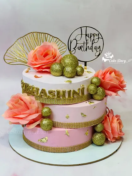 An elegant three-tier cake in shades of pink and gold. This crisscross design is adorned with sparkling gold balls and soft peach roses, perfect for a grand birthday celebration.