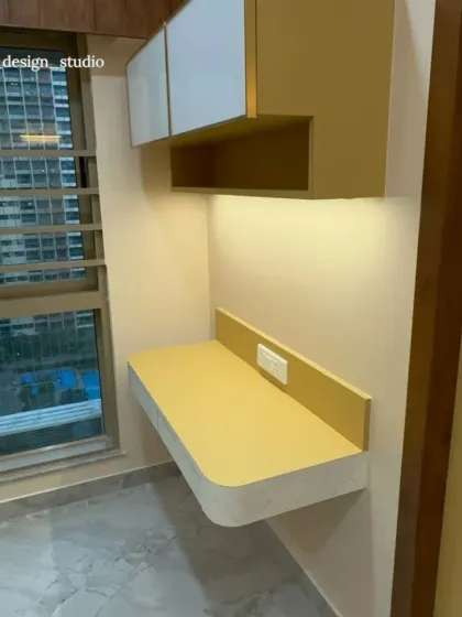 A minimalist and modern study nook designed for focus. The floating desk in a cheerful mustard yellow is paired with clean white overhead cabinets, creating a bright and uncluttered workspace that gets plenty of natural light from the adjacent window.