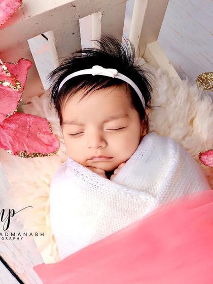 A baby girl sleeping in a white bed, surrounded by large, glittery pink flowers.