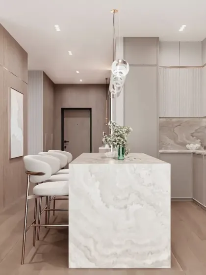 A luxurious open-plan kitchen with a large marble island and bar seating. The light wood and beige tones create a warm and sophisticated atmosphere.