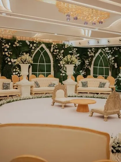 A serene and elegant indoor mandap setup. The circular white stage is surrounded by plush white sofas against a wall of green foliage and white floral accents, creating a feeling of a secret garden indoors.