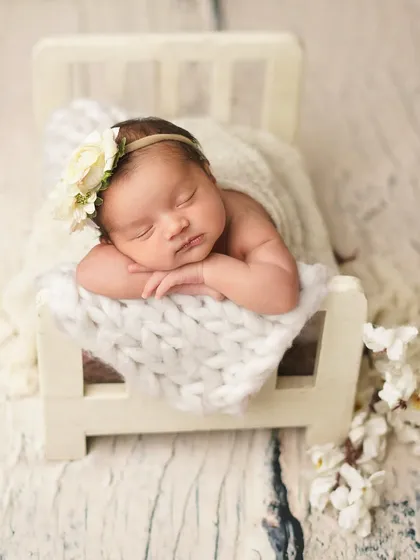 This little one is dreaming sweetly, resting her head on her hands in a tiny bed. The floral headband and white blossoms add a touch of delicate, natural beauty to the infant photoshoot.