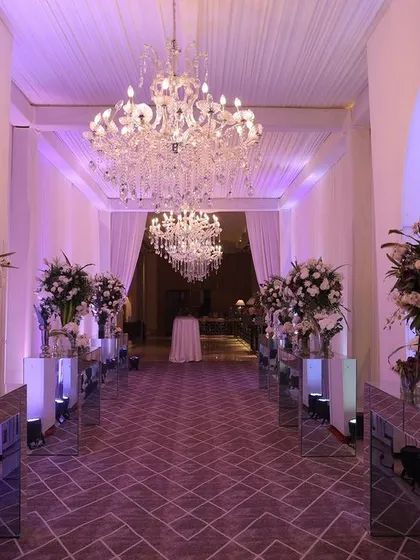 A grand and dramatic entrance for a reception at The Oberoi, Gurgaon. The passage is lined with large white floral arrangements on mirrored pedestals, all under the glow of magnificent crystal chandeliers.