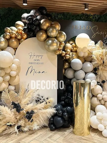 A luxurious 60th birthday decoration with a 'Hello Sixty' theme. The setup includes a personalized backdrop, a rich arrangement of black, nude, and gold balloons, and dried pampas grass for texture.