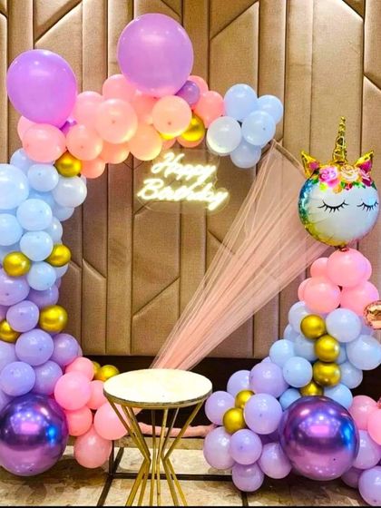 A magical unicorn-themed birthday decoration featuring a large unicorn foil balloon and a pastel balloon arch. The setup includes a 'Happy Birthday' neon sign and a number '7' foil balloon to mark the special age.