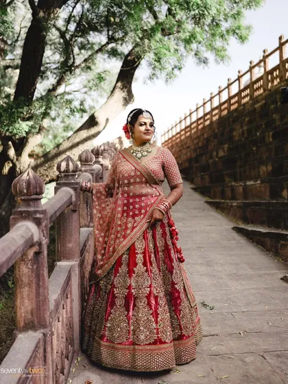 A stunning portrait of a bride in her red lehenga, standing on a stone walkway, the intricate details of her outfit shining in the soft light.