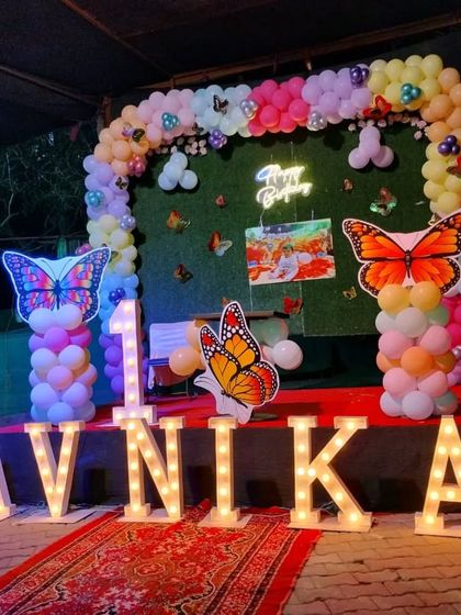 A vibrant butterfly-themed outdoor party with large marquee letters spelling 'AVNIKA'. The setup includes a grass wall backdrop, colorful balloon columns with butterfly cutouts, and a light-up number '1'.