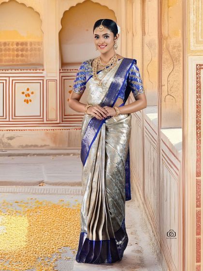 A full-length shot of a model in a magnificent silver and blue silk saree. The grand, traditional setting and beautiful lighting create a regal and timeless bridal portrait.