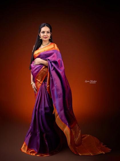 A standing portrait showcasing the beautiful drape of a traditional silk saree. The rich purple and orange colors look stunning against the warm, glowing backdrop.