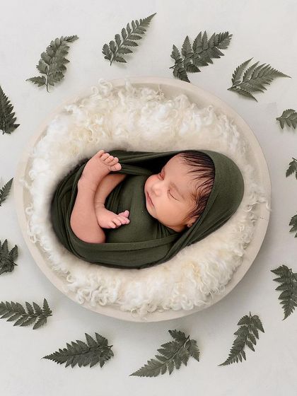 A minimalist and artistic top-down shot of a baby swaddled in green, lying in a white bowl and encircled by delicate fern leaves.