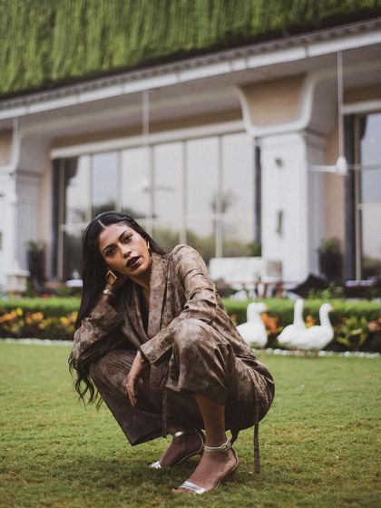 Anasuya Sengupta crouching in the grass, looking directly at the camera. Her pose is strong and grounded, connecting her with the natural surroundings.
