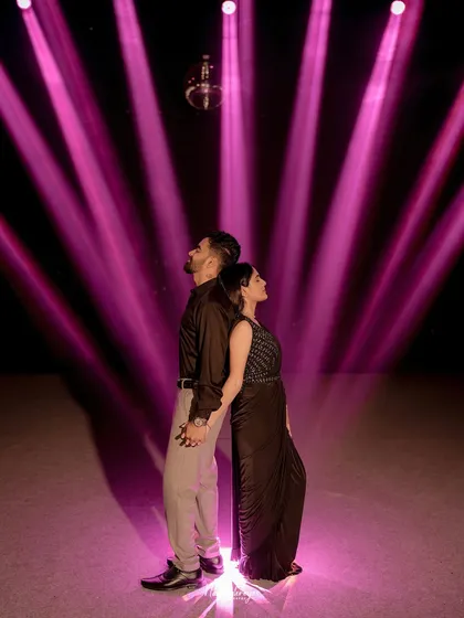 A stylish shot of a couple standing back-to-back under a disco ball and dramatic pink spotlights, creating a fun and energetic party vibe.