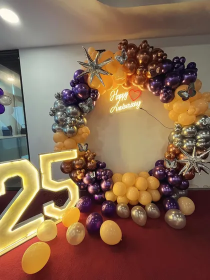 A stylish ring decoration for a 25th anniversary, using purple, orange, and chrome balloons with star and butterfly accents. It also features a 'Happy Anniversary' neon sign and large marquee numbers.
