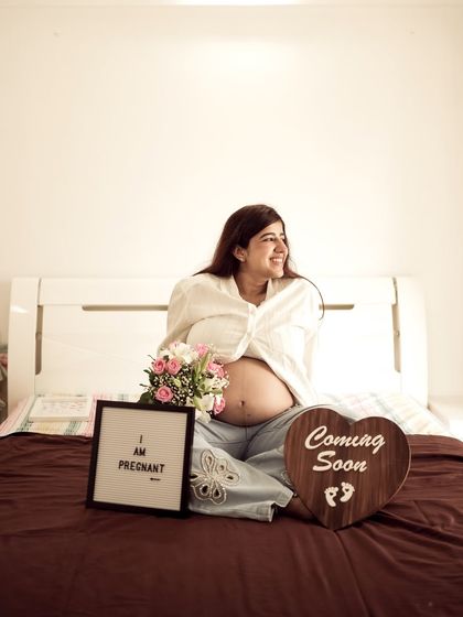 A joyful indoor portrait of the mom-to-be sitting on a bed, surrounded by flowers and "I am Pregnant" and "Coming Soon" signs.