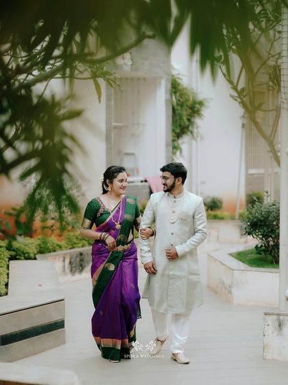 The couple walking together through a modern courtyard, the bride's vibrant purple saree standing out beautifully.