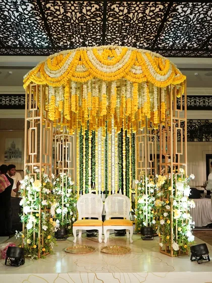 A beautiful Haldi seating area, where the couple is framed by a modern golden structure adorned with a canopy of yellow and white marigold garlands.