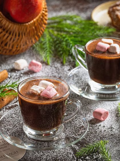 A cozy winter scene with two mugs of hot chocolate topped with marshmallows, styled with cinnamon sticks and pine branches.
