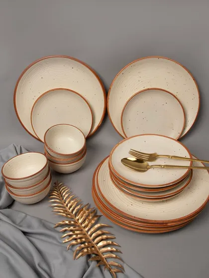 A complete cream-colored, speckled ceramic dinnerware set, elegantly arranged with a golden leaf prop and grey fabric.