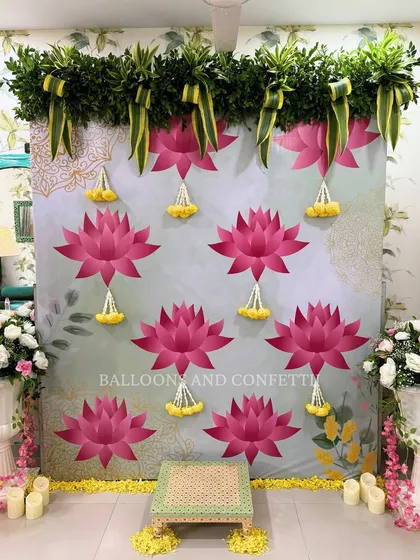 This is a closer look at a vintage-inspired puja backdrop. The design features a custom printed cloth with large pink lotus motifs, topped with a fresh green foliage border. The hanging floral tassels and the traditional bajot for the ceremony complete this serene and elegant setup.