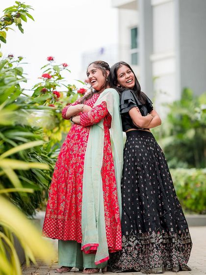 Two sisters laughing and posing back-to-back in a garden. Their coordinated outfits and joyful expressions make this a perfect sibling portrait.