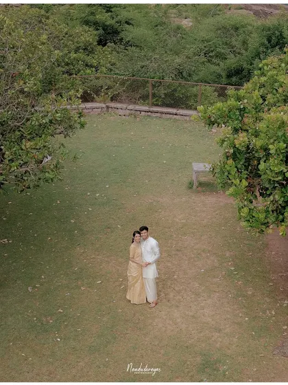 A drone shot of a couple standing together in a green clearing, showcasing the beautiful landscape surrounding them.