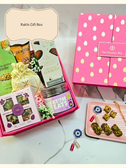 A beautiful shot of the pink Rakhi Gift Box, filled with treats like cookies, brittle, and dry fruit bites. It comes with a traditional Rakhi and a protective Evil Eye Roli Chawal set.
