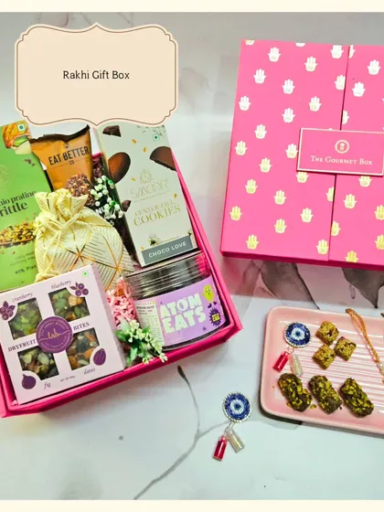 A beautiful shot of the pink Rakhi Gift Box, filled with treats like cookies, brittle, and dry fruit bites. It comes with a traditional Rakhi and a protective Evil Eye Roli Chawal set.