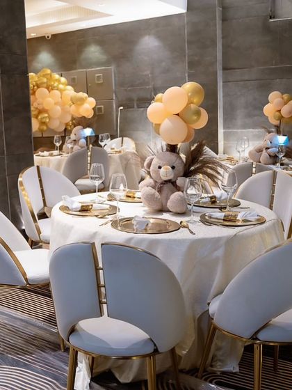 Elegant table settings for a teddy bear themed birthday. Each table has a plush bear centerpiece with balloons, complemented by gold charger plates and modern white chairs.