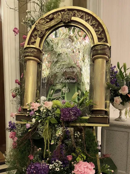 A welcome sign set in an ornate, antique-gold mirrored frame, surrounded by lush purple and pink florals, creating a grand and artistic entrance.
